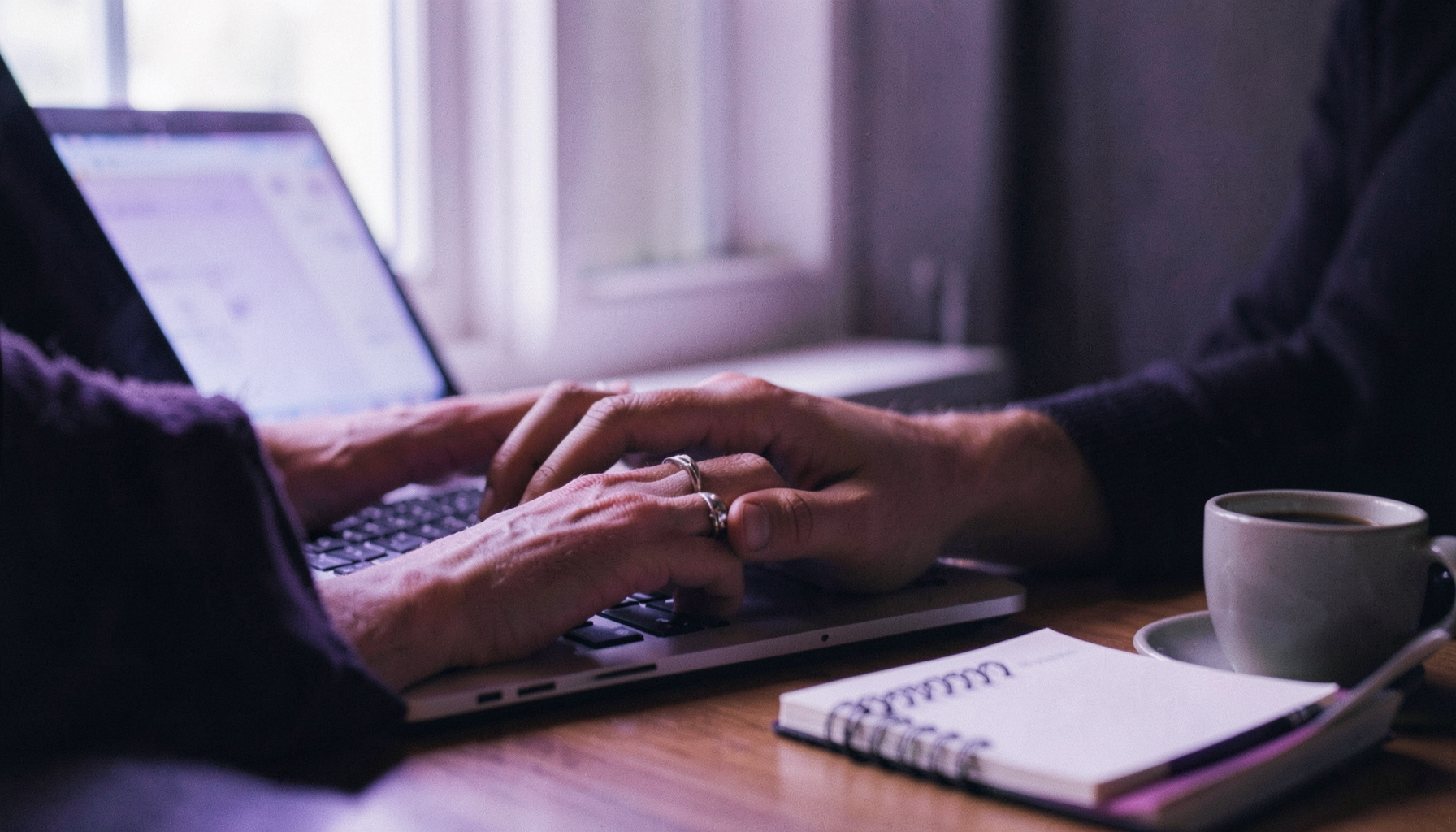 A close-up of hands typing on a laptop with a notebook and coffee nearby, soft natural light from a window, evoking focused remote collaboration.